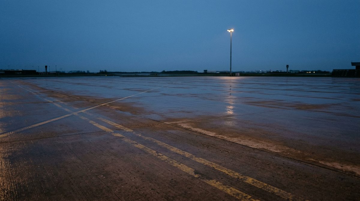 A wide empty concrete apron at dusk under a deep blue sky, wet concrete reflecting a single distant floodlight