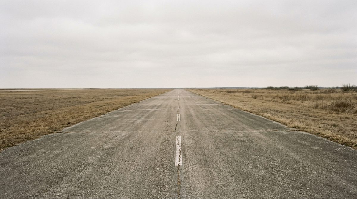 An empty runway on flat open prairie seen straight down its center line, dormant tan grass to either side under a wide pale sky